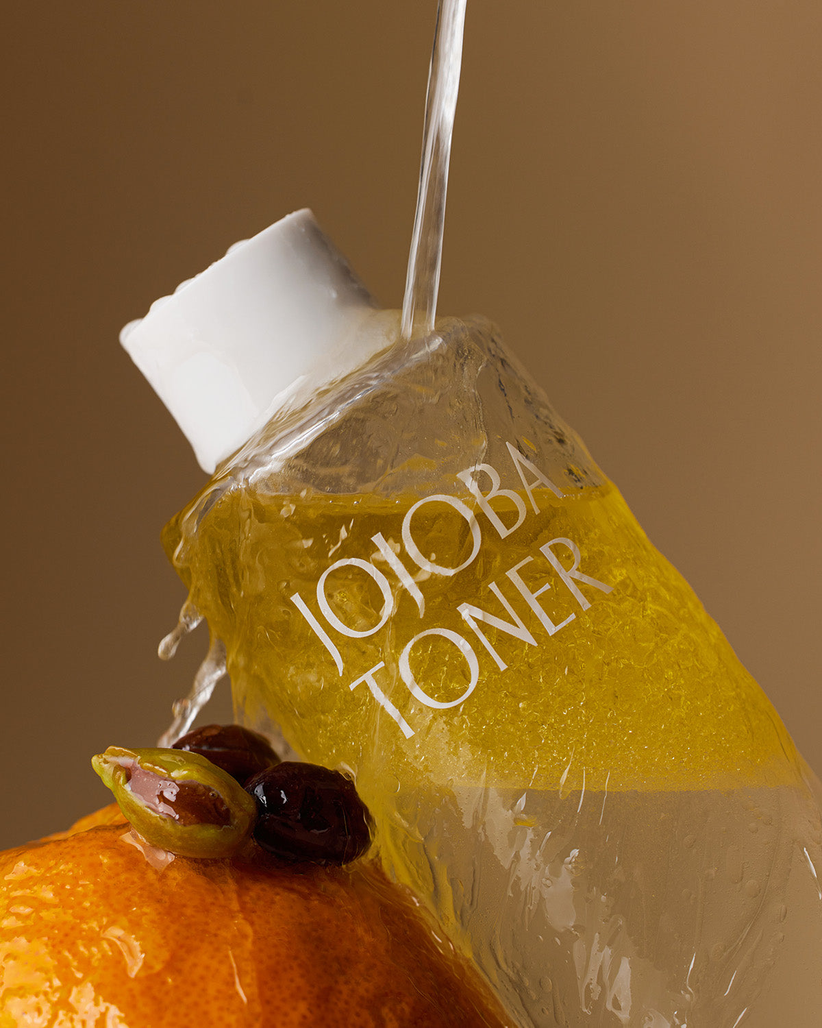 A dramatic close-up of the Jojoba Toner bottle on its side, with water pouring over the cap and bottle. The bottle rests on an orange fruit, and a few jojoba seeds are visible in the foreground.