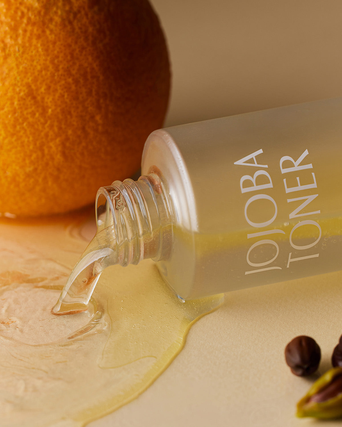 A close-up, artistic shot of the Jojoba Toner bottle lying on its side, with the yellow-tinted, oily toner spilling out onto a tan surface. In the background are a large orange fruit and a few jojoba seeds.