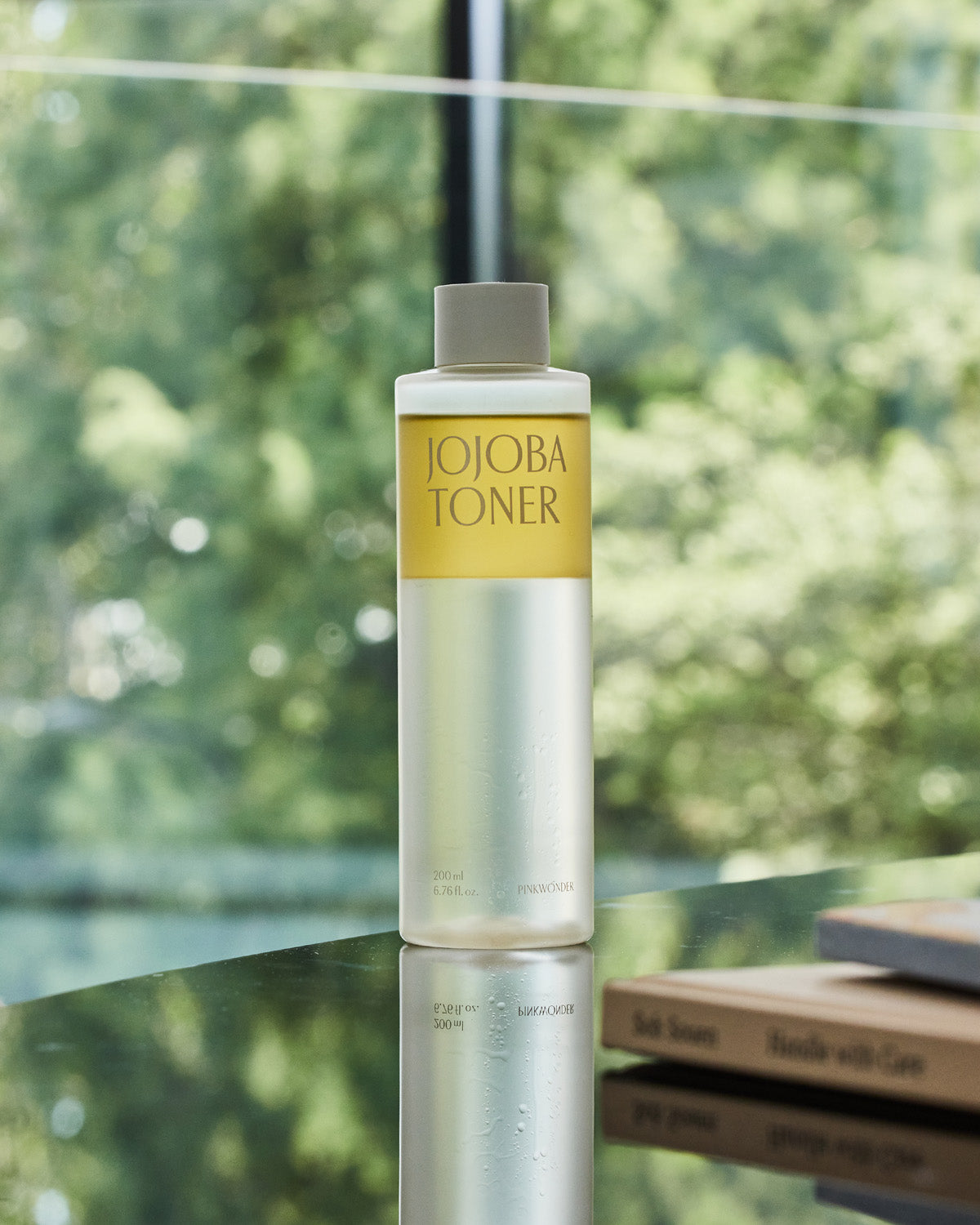 A clear glass bottle of Pinkwonder Jojoba Toner with a distinctive yellow and clear liquid separation stands on a reflective surface against a blurred green outdoor background seen through a window. The cap is a light gray-beige.