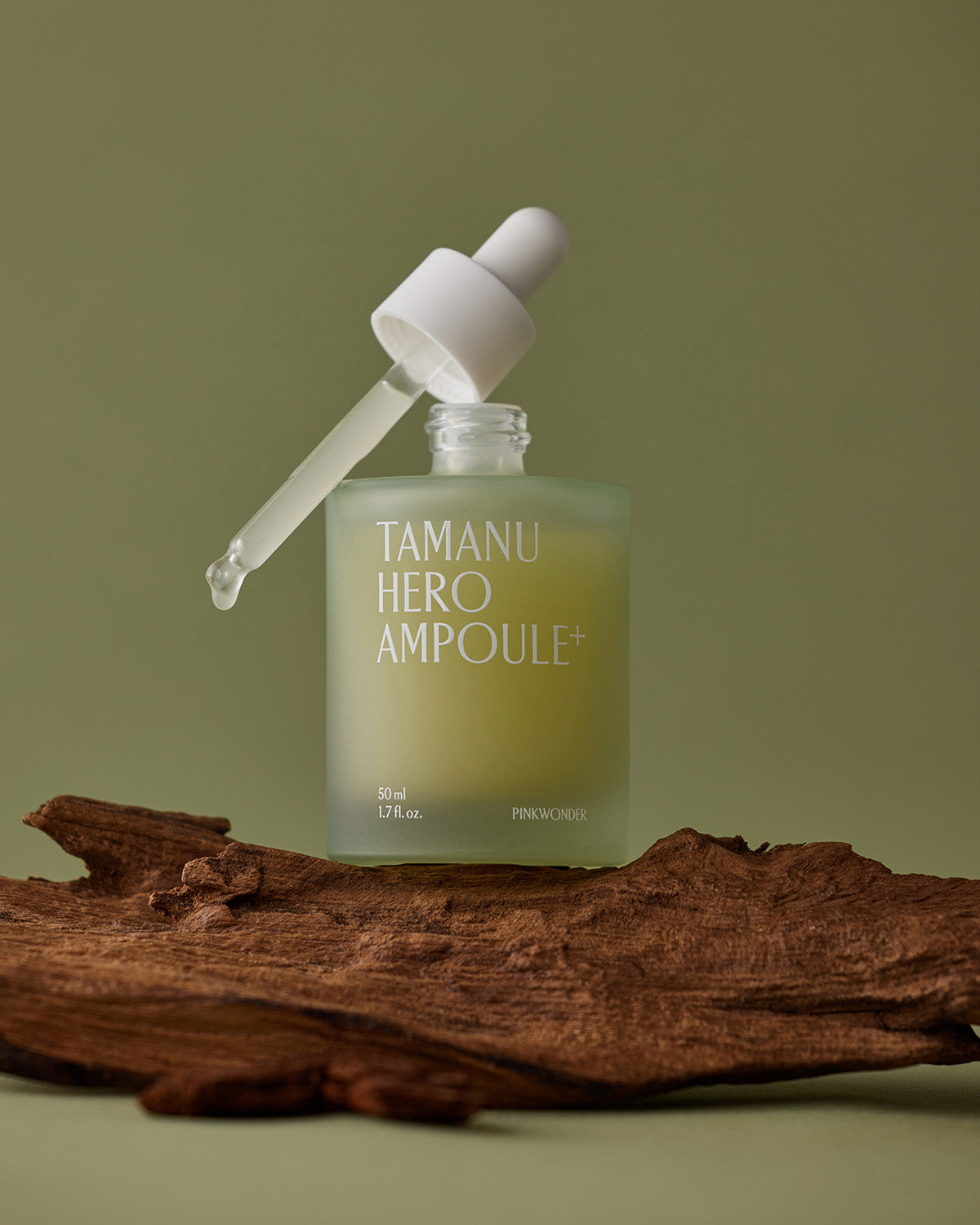 The Tamanu Hero Ampoule+ bottle rests on a piece of dark, textured wood against a pale green background. The glass dropper is suspended over the bottle, dropping a bead of serum.