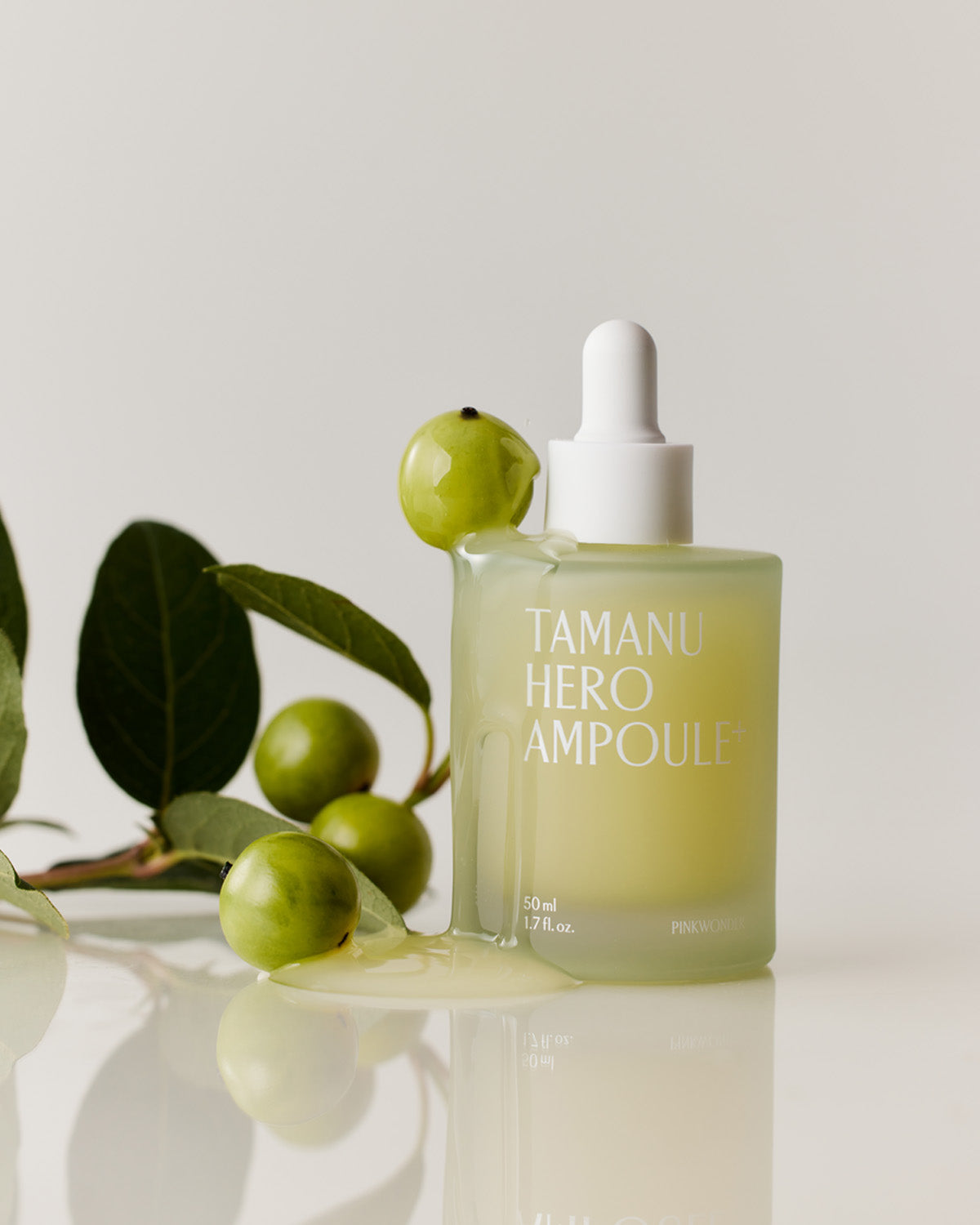 The Tamanu Hero Ampoule+ bottle stands on a white reflective surface next to a leafy branch with green, round fruit, emphasizing the tamanu ingredient.