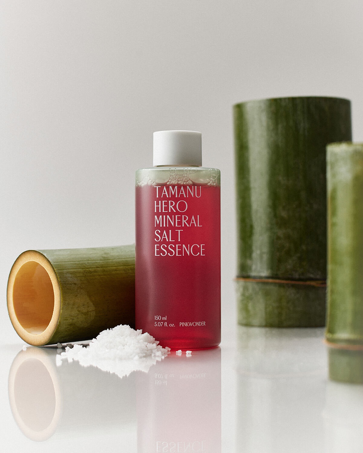 A bottle of the pink-red Tamanu Hero Mineral Salt Essence sits on a reflective white surface next to scattered white salt crystals and pieces of green bamboo.
