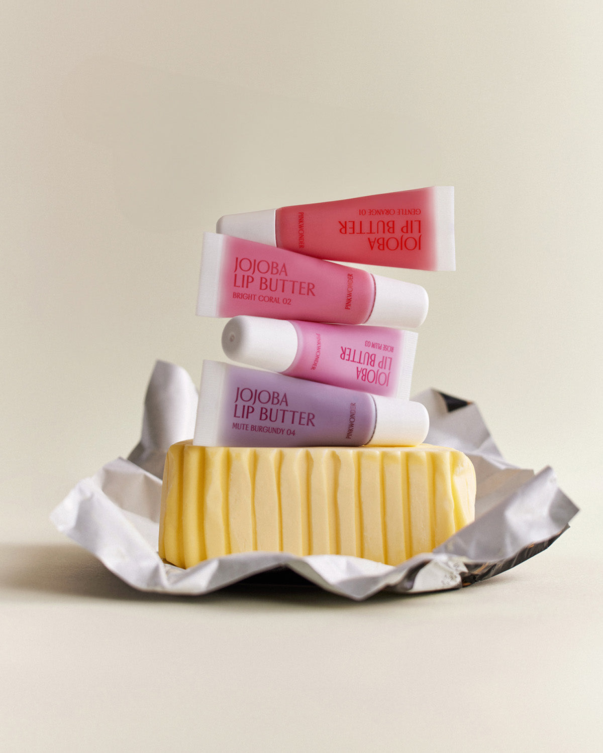 An artistic still life showing four tubes of the colored Jojoba Lip Butter stacked on top of a rectangular stick of butter that is wrapped in crumpled foil, playing on the "lip butter" name.