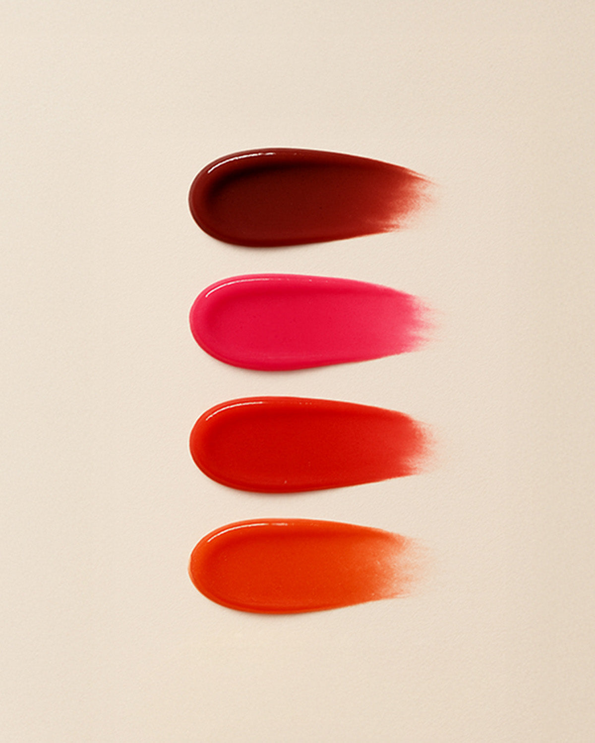 A flat-lay photo of four glossy lip swatches on a pale tan background, demonstrating the shade range. From top to bottom: a deep burgundy, a bright magenta, a true red-pink, and a warm coral-orange.