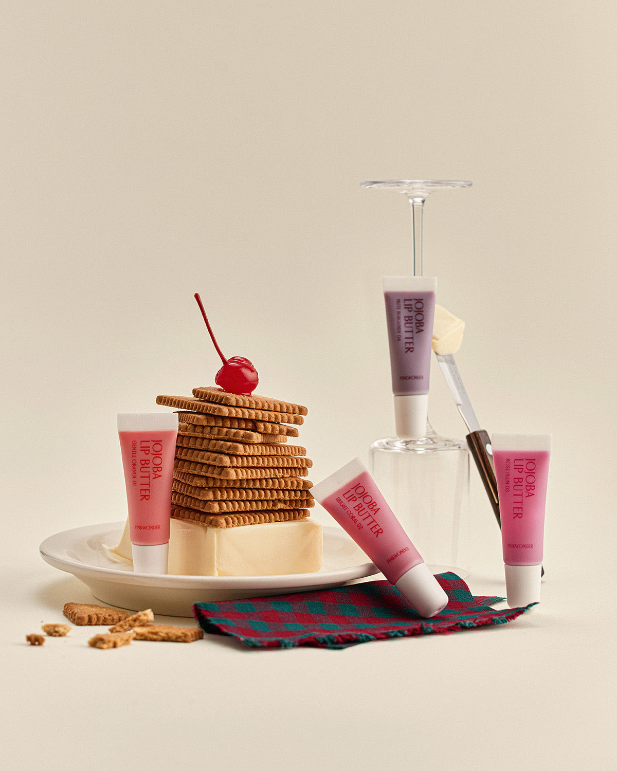 A playful still life featuring stacks of cookies and a stick of butter on a plate, topped with a maraschino cherry. Four tubes of Jojoba Lip Butter are artfully placed around the food, using upturned glassware as props.