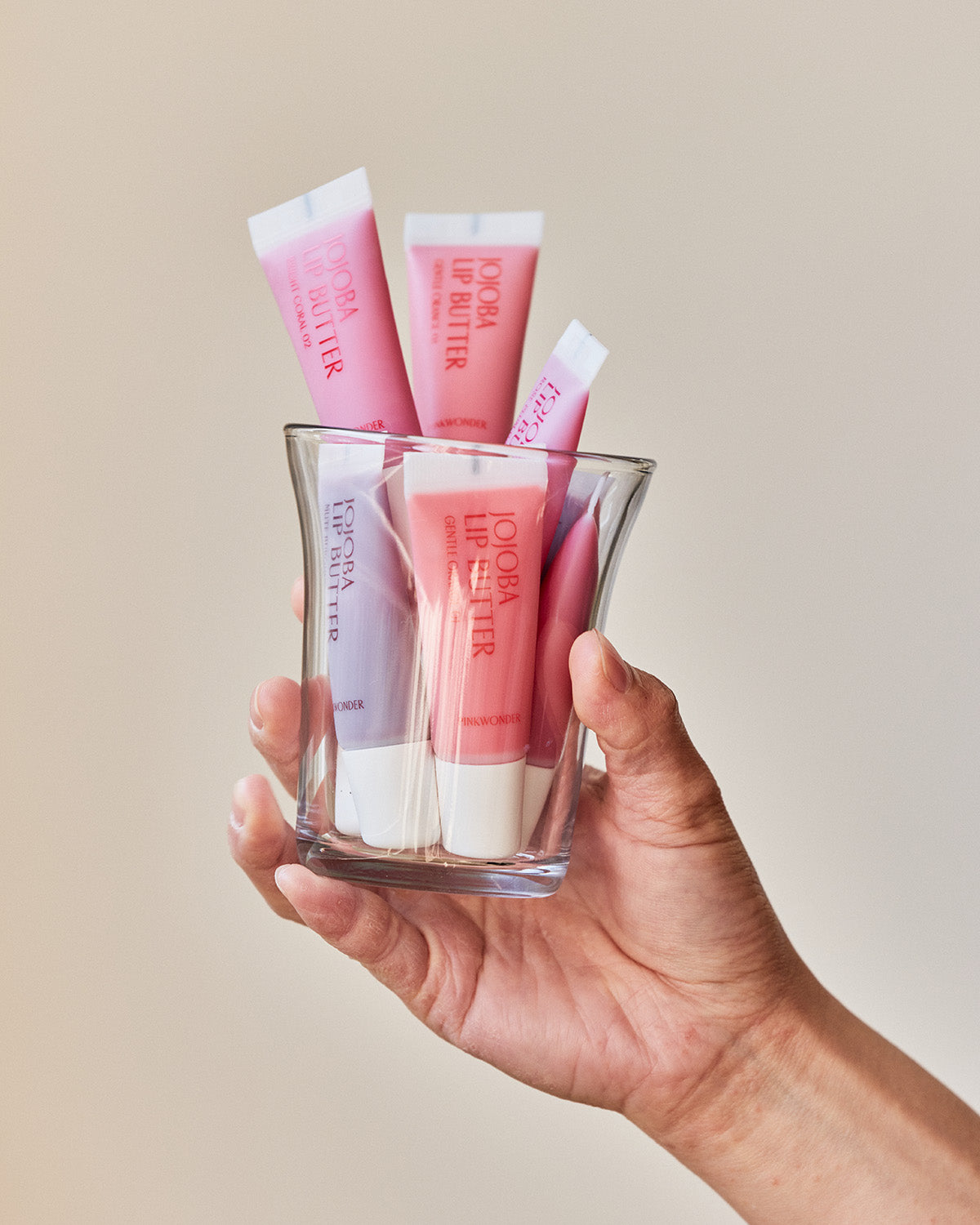 A hand holds a clear glass filled with various tubes of the Pinkwonder Jojoba Lip Butter in different shades, set against a simple, light-colored background.