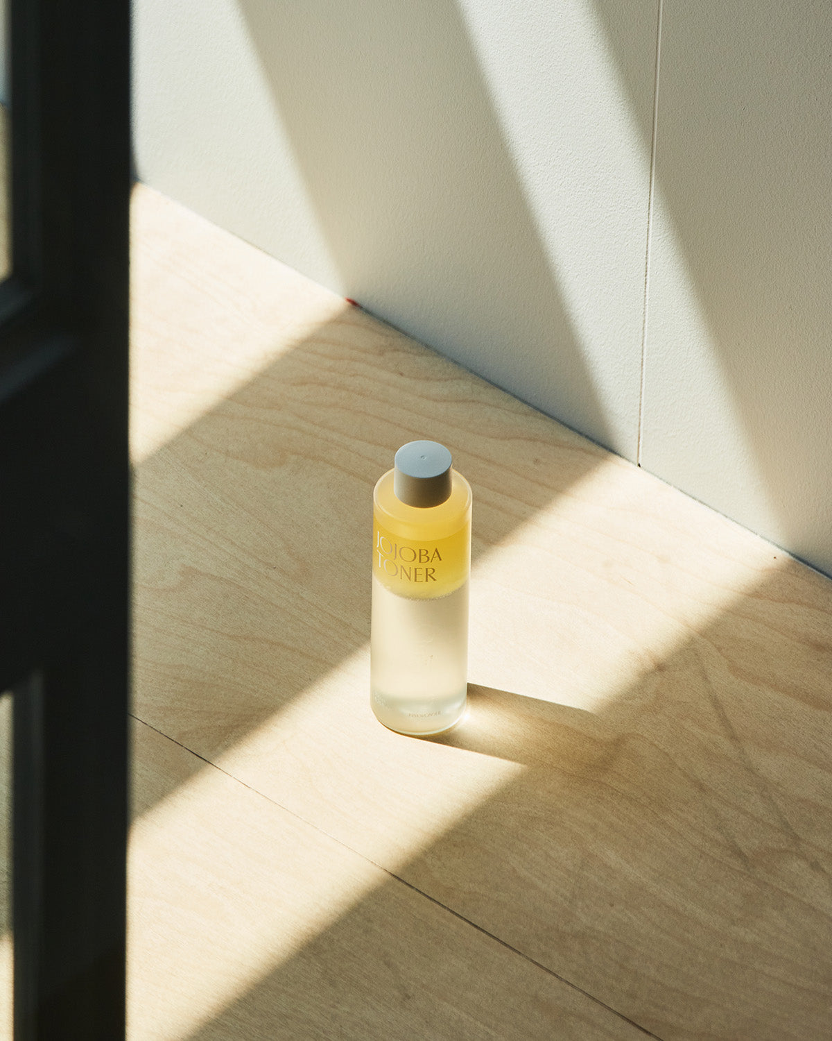 A minimalist product shot of the Jojoba Toner bottle standing on a light wooden floor with a white wall behind it, bathed in a strong, angular beam of natural sunlight creating striking shadows.