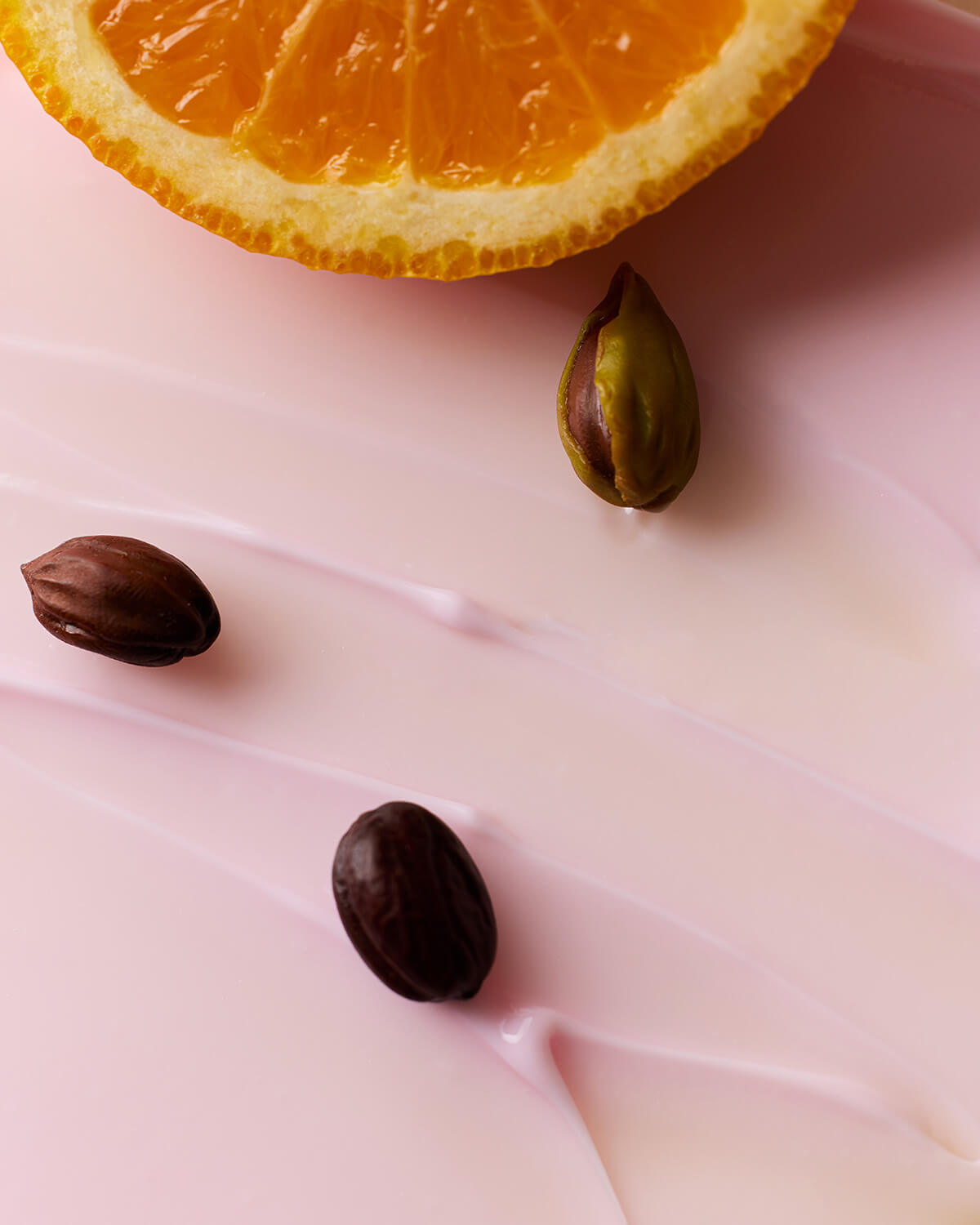 A close-up texture shot of pale pink cream swiped across a surface. Three brown and green jojoba seeds rest on the cream, and a slice of orange is visible along the top edge of the frame, emphasizing the vitamin ingredients.