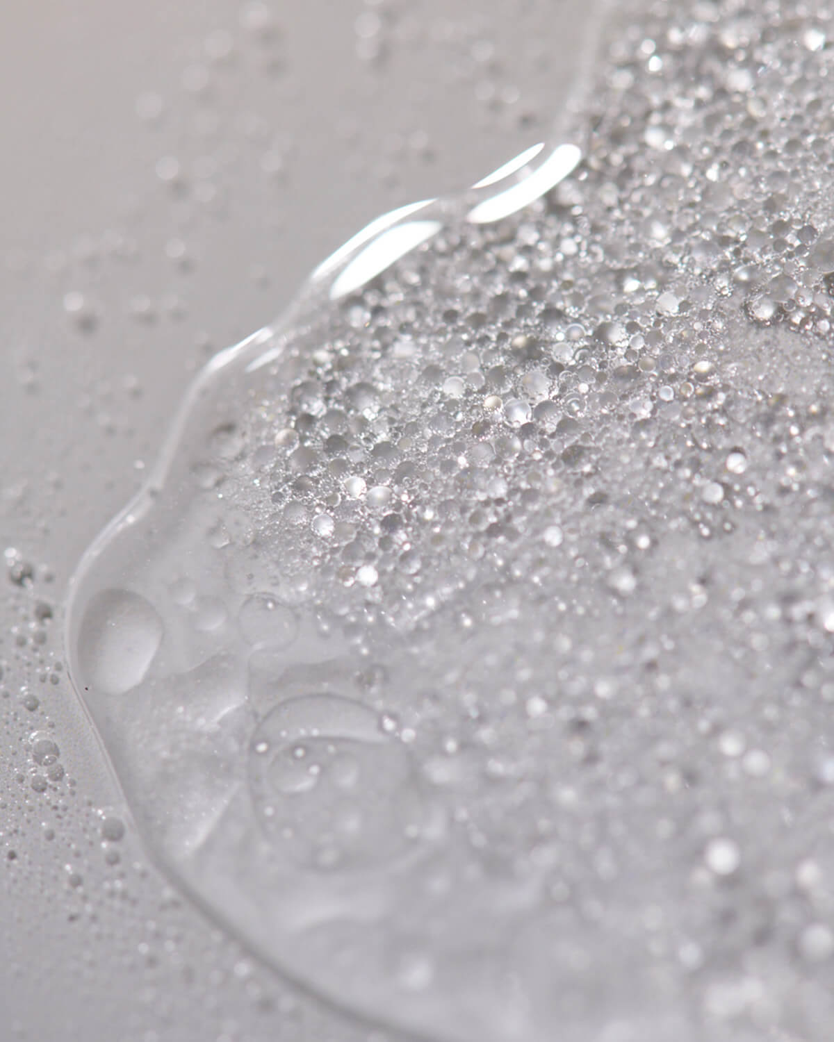 An abstract, macro shot of a clear liquid, possibly the mist, spilled on a light gray surface, with large and small shimmering bubbles creating a textured pattern.