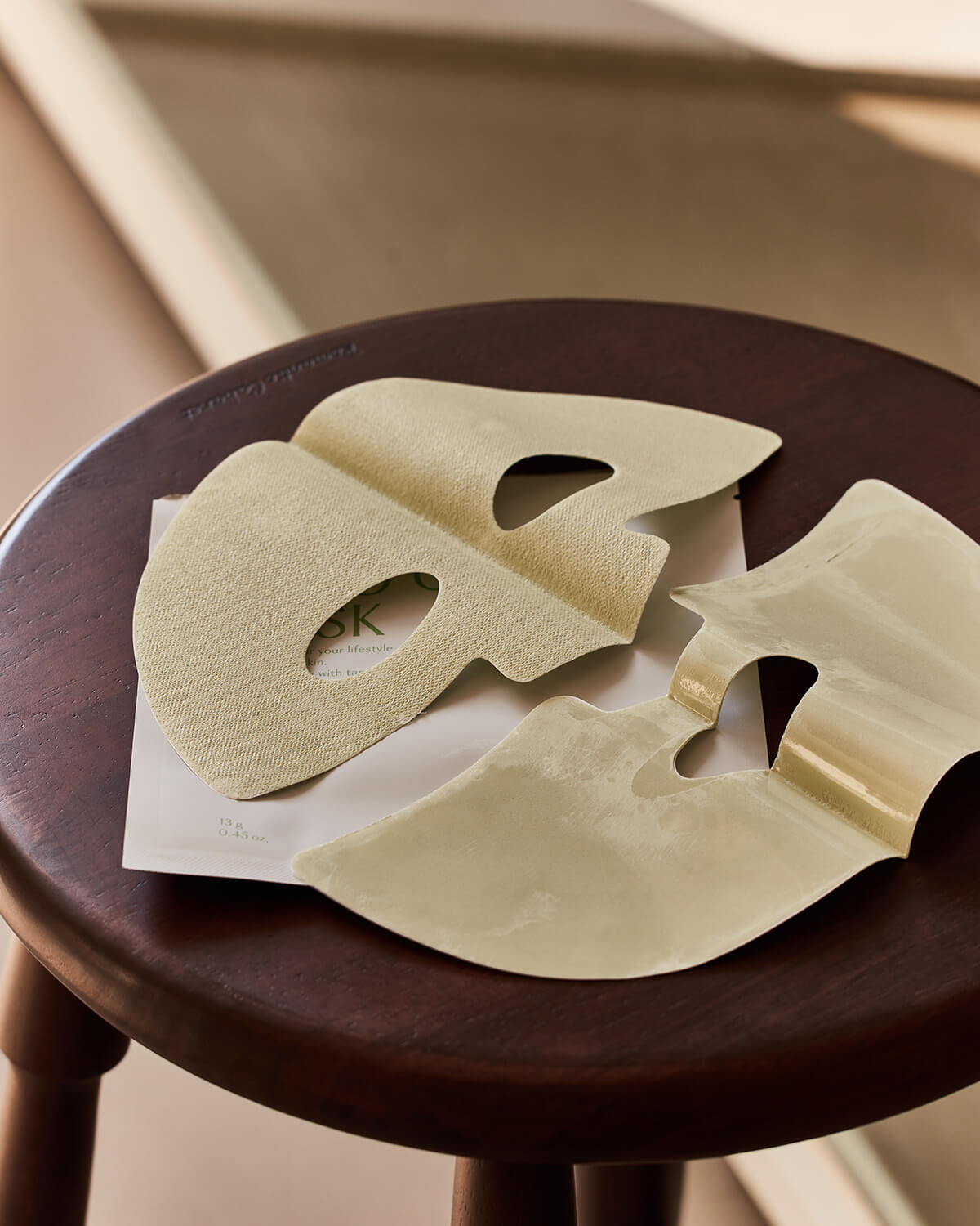 A new, two-piece, light tan-colored clay sheet mask is spread open on a small, dark wooden stool.