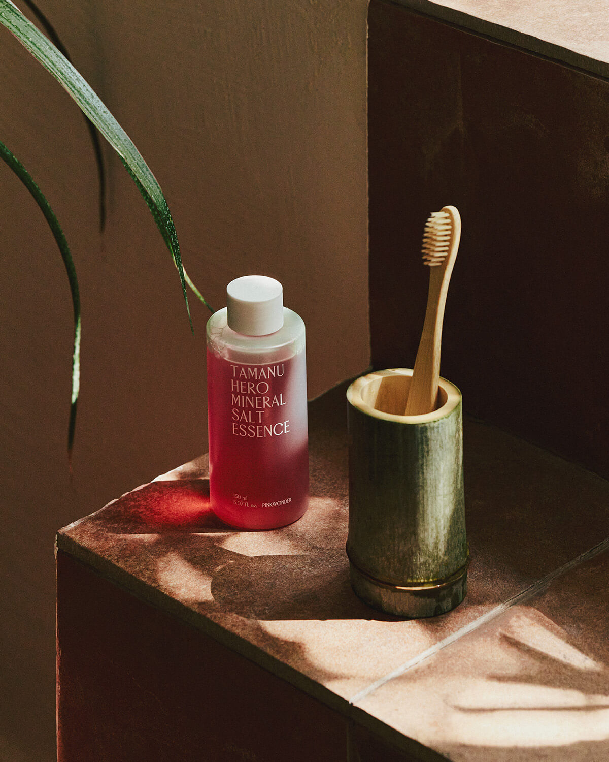 The bottle of pink-red Tamanu Hero Mineral Salt Essence sits on a brown tiled surface next to a wooden toothbrush in a bamboo cup holder.