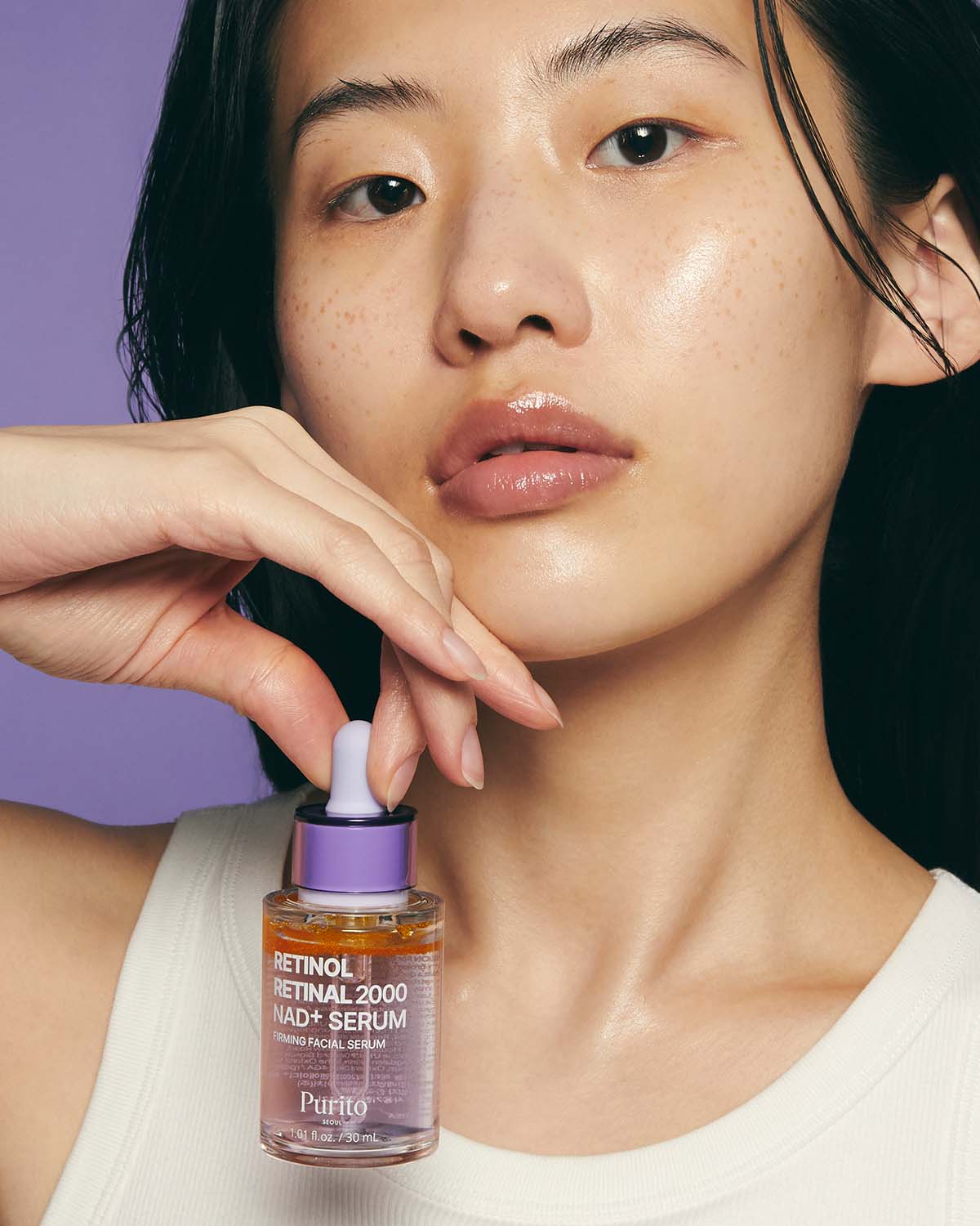 Close-up of a model with glowing skin holding the Purito Retinol Retinal 2000 NAD+ Serum bottle.