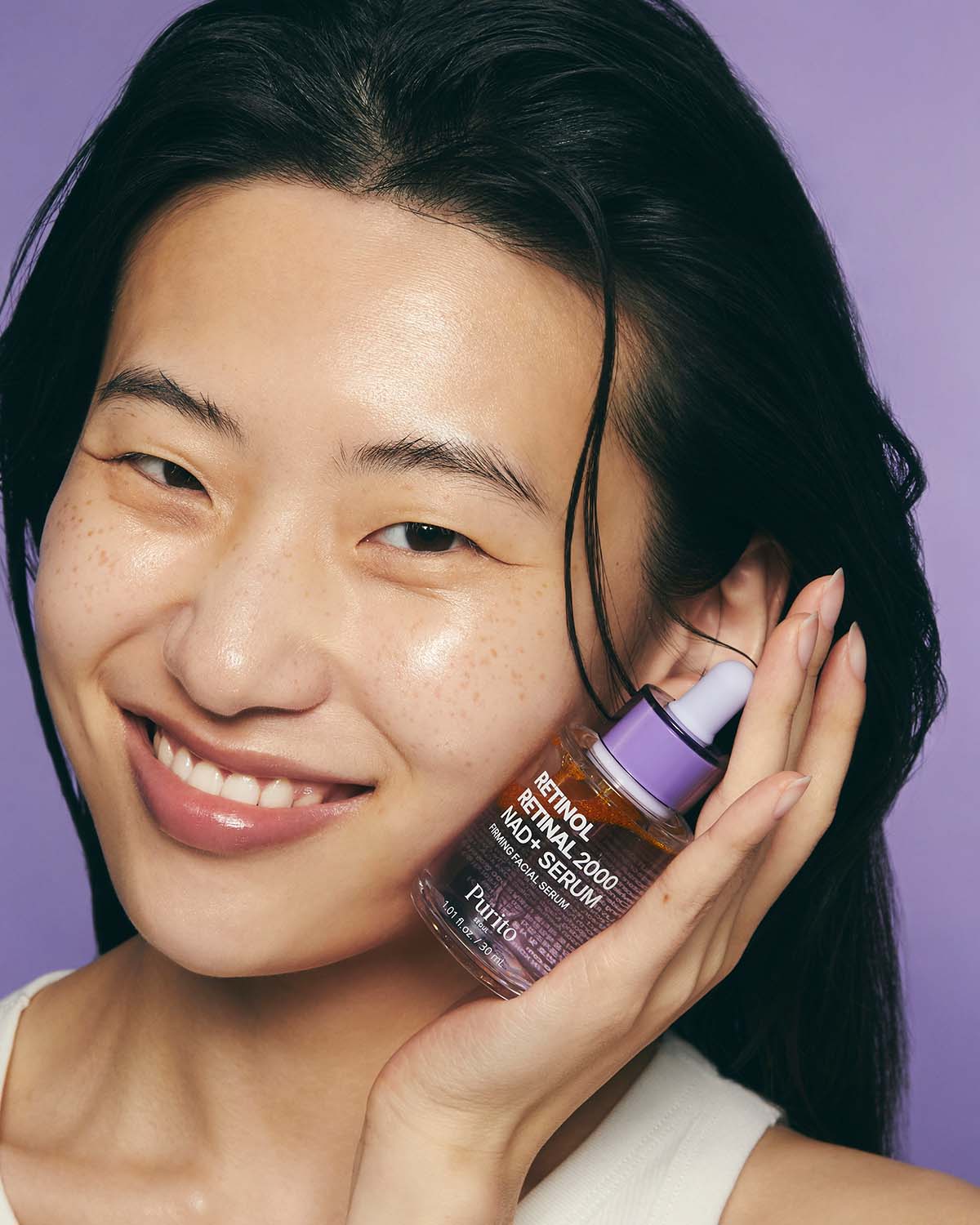 Smiling model holding the Purito Retinol Retinal 2000 NAD+ Serum bottle next to her face.
