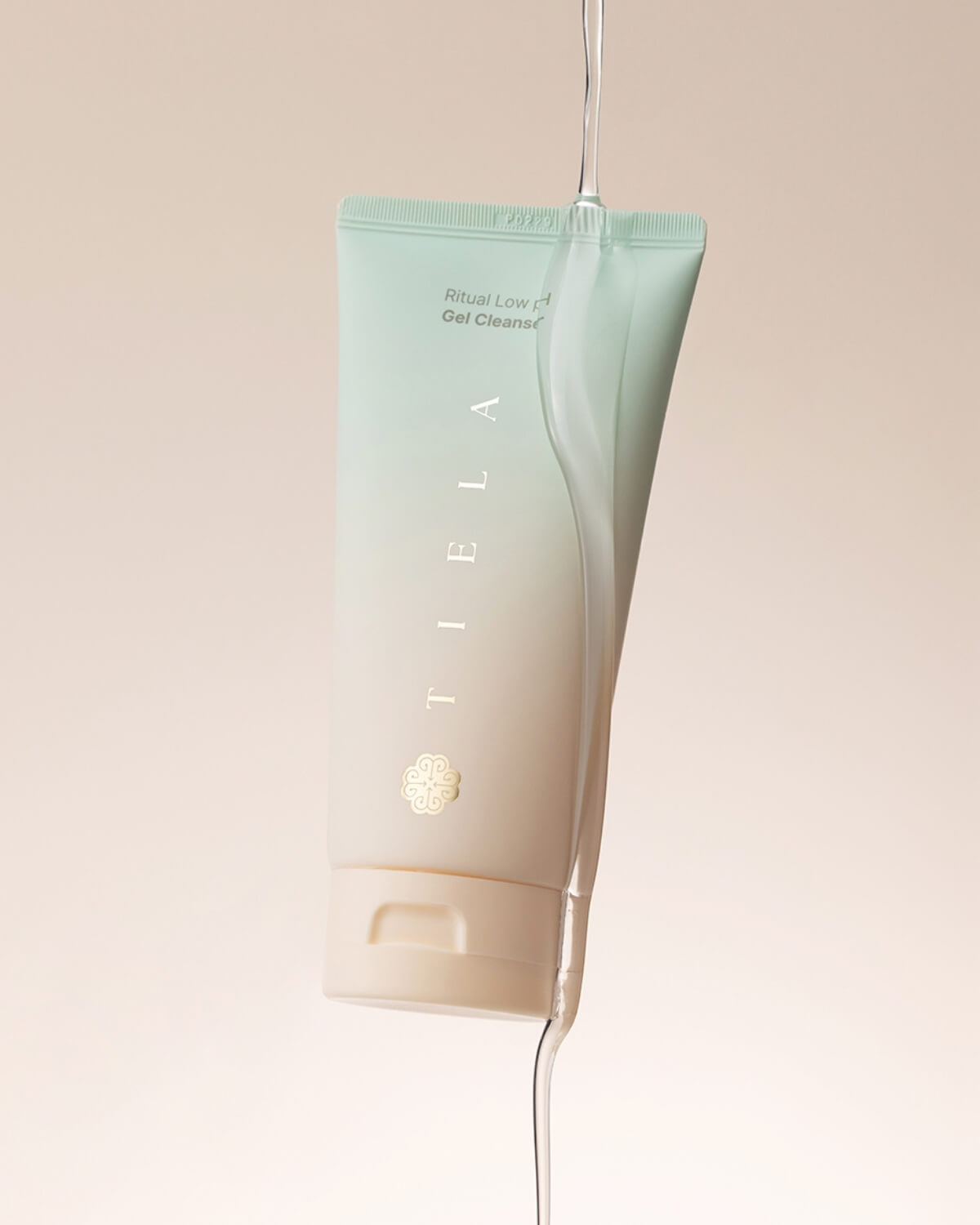 The Tiela Low pH Gel Cleanser tube is suspended in the air, with clear liquid pouring over and around it, suggesting a cleansing or watery texture.