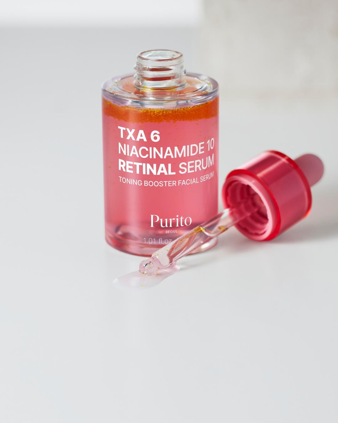 A close-up shot of the Purito TXA 6 Niacinamide 10 Retinal Serum with the dropper resting outside the bottle on a white surface, where a small amount of the serum has dripped. The bottle shows the separated pink and orange bi-phase serum.