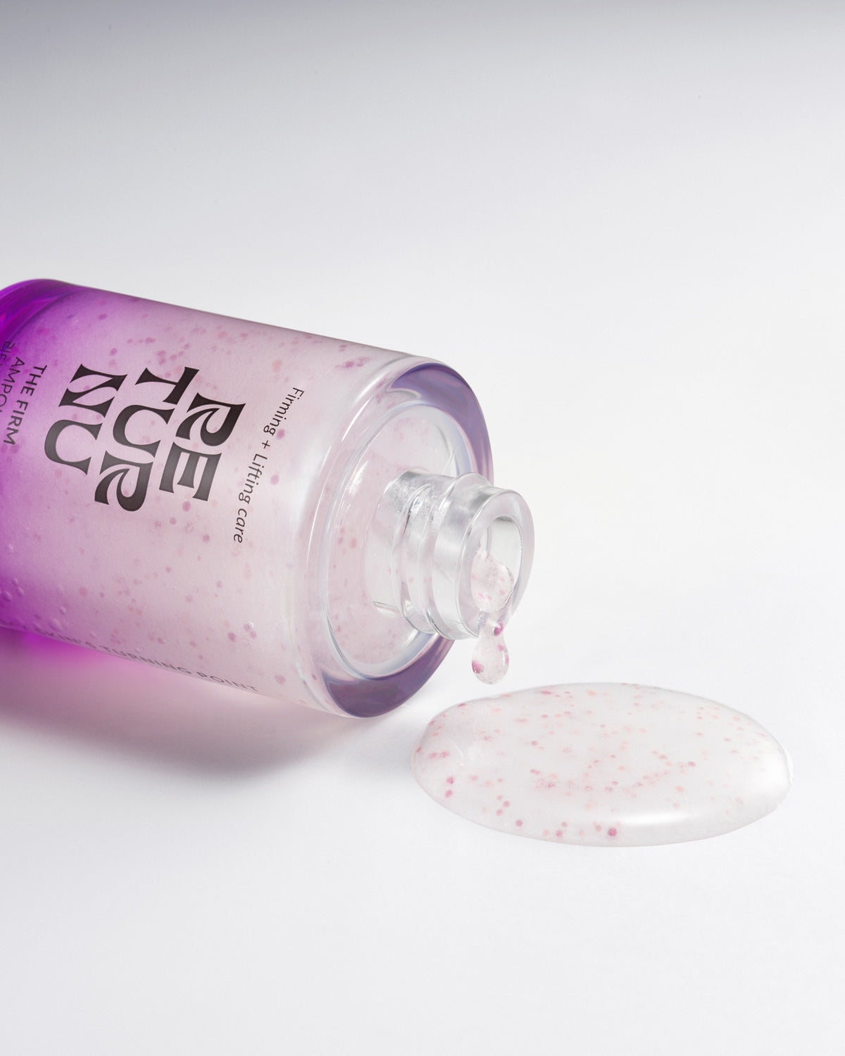The ampoule bottle tipped on its side with a puddle of clear serum containing small pink capsules on a white surface.