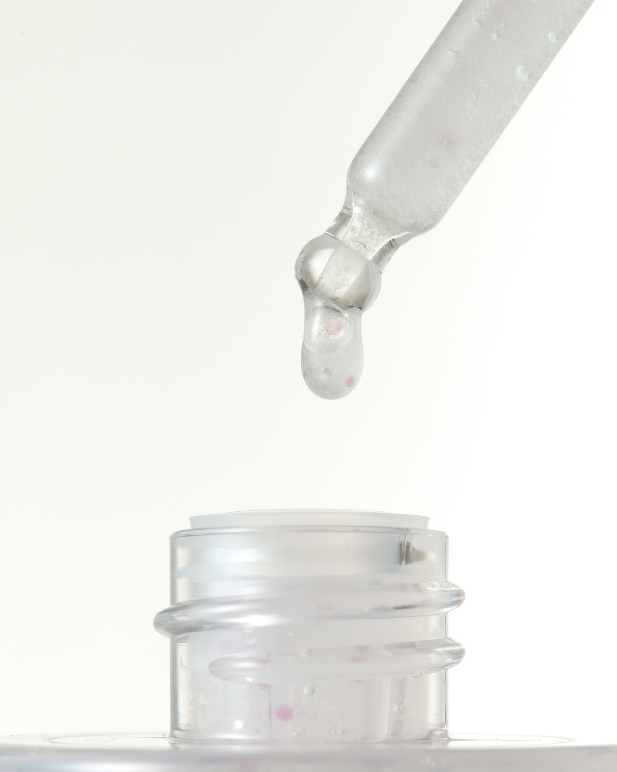 A macro shot of a glass dropper dispensing a single drop of clear serum with pink capsules back into the bottle opening.