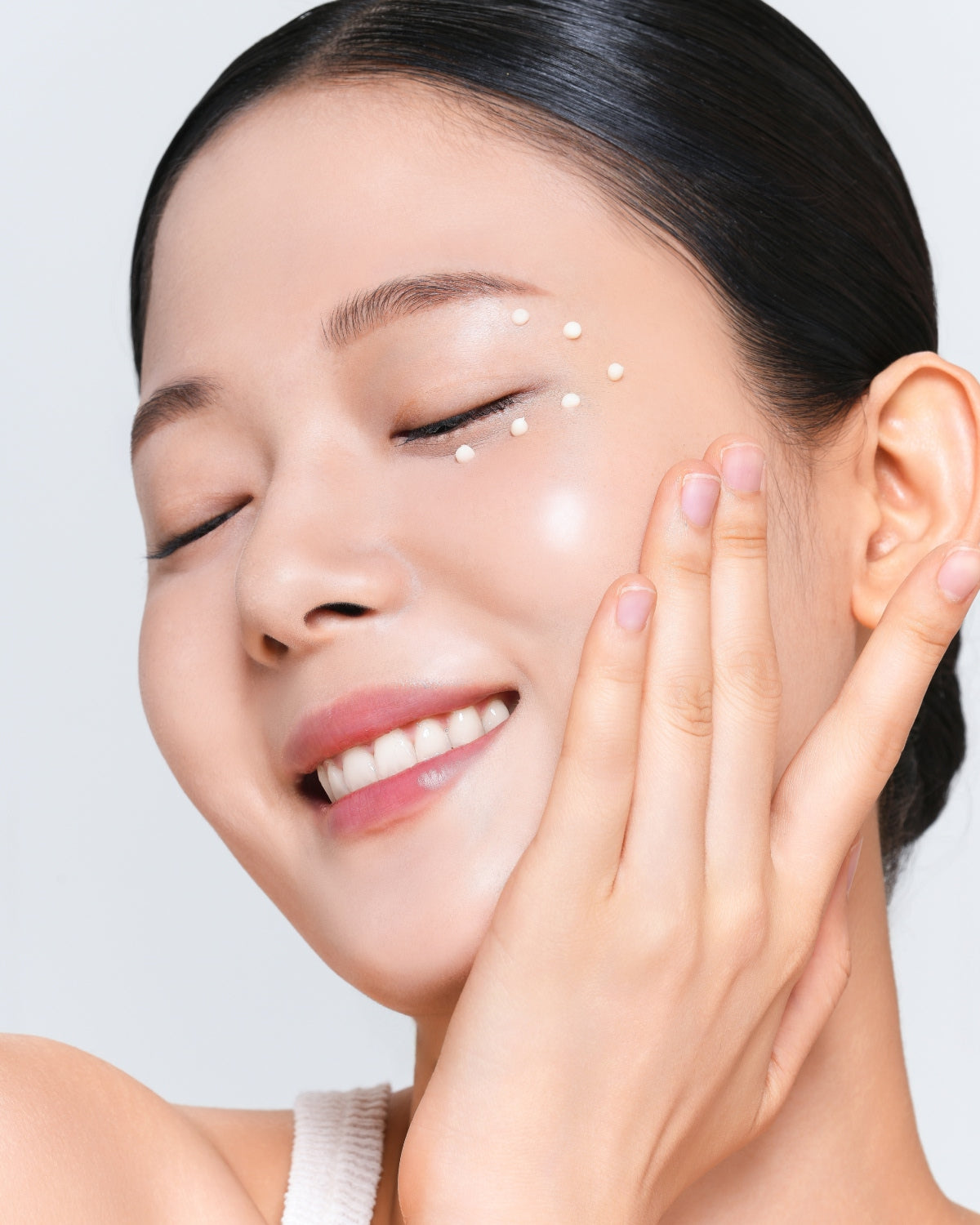 A close-up of a person smiling with eyes closed, featuring small dots of cream applied around the outer corner of their eye.