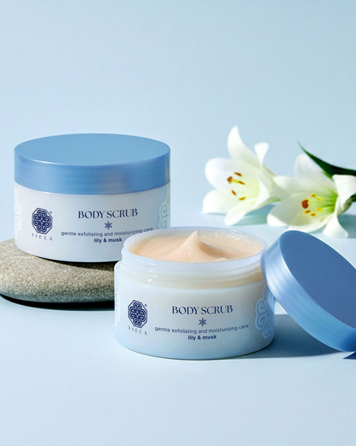 Two jars of Tiela Lily & Musk body scrub on a stone with white lilies against a soft blue background.