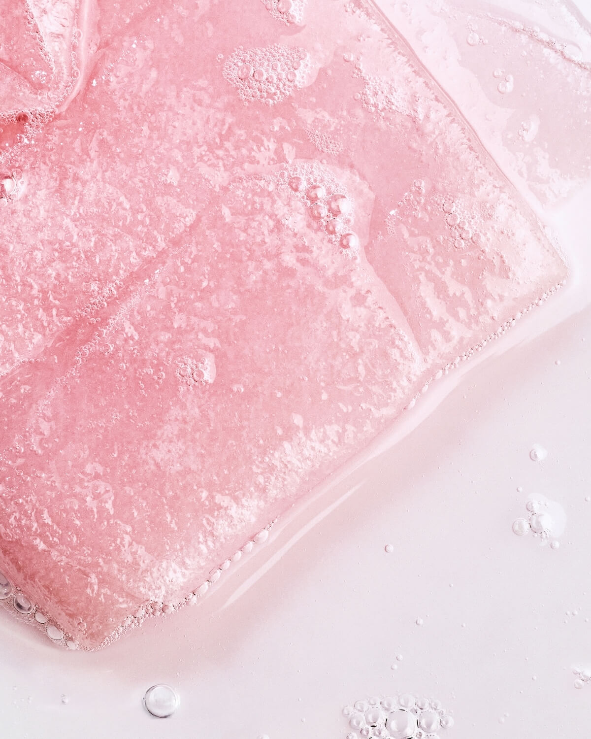 Abstract close-up of the pink sheet mask submerged in liquid, showing bubbles and a glossy, wet texture.