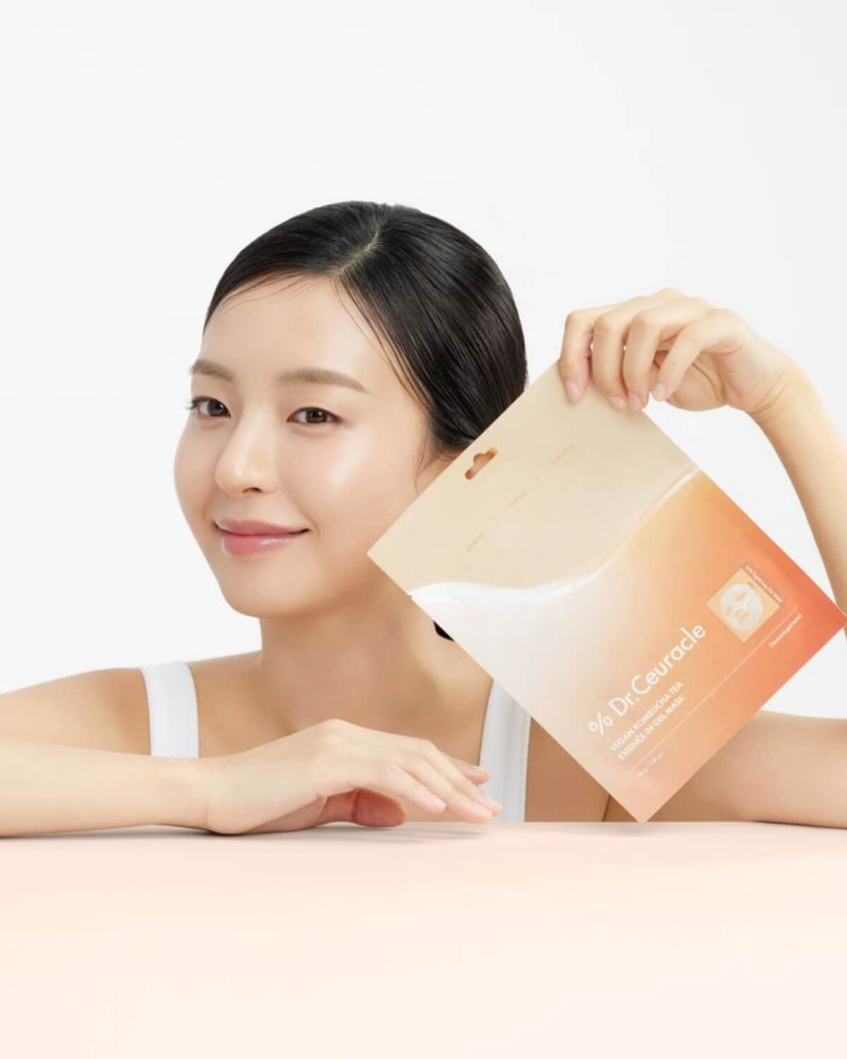 A woman smiling while holding the Dr. Ceuracle Vegan Kombucha Tea Essence in Gel Mask pouch against a neutral background.