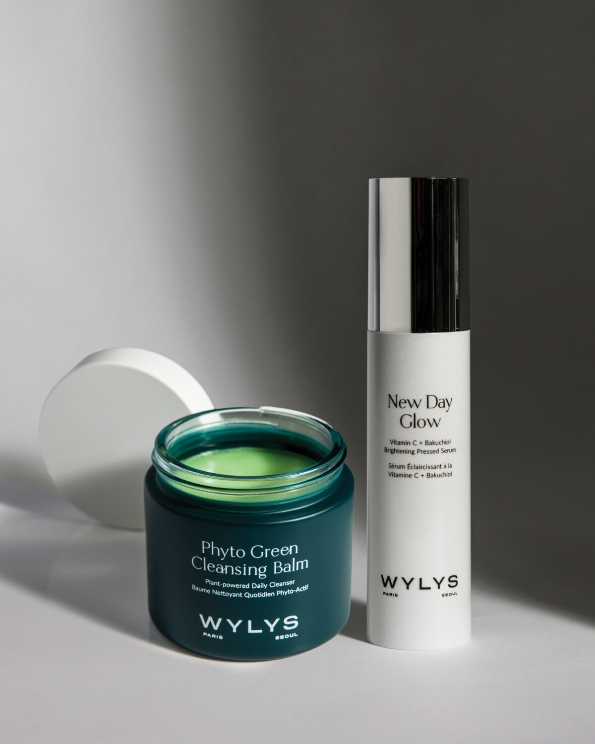 WYLYS Phyto Green Cleansing Balm and New Day Glow Brightening Pressed Serum on a white surface with shadows.