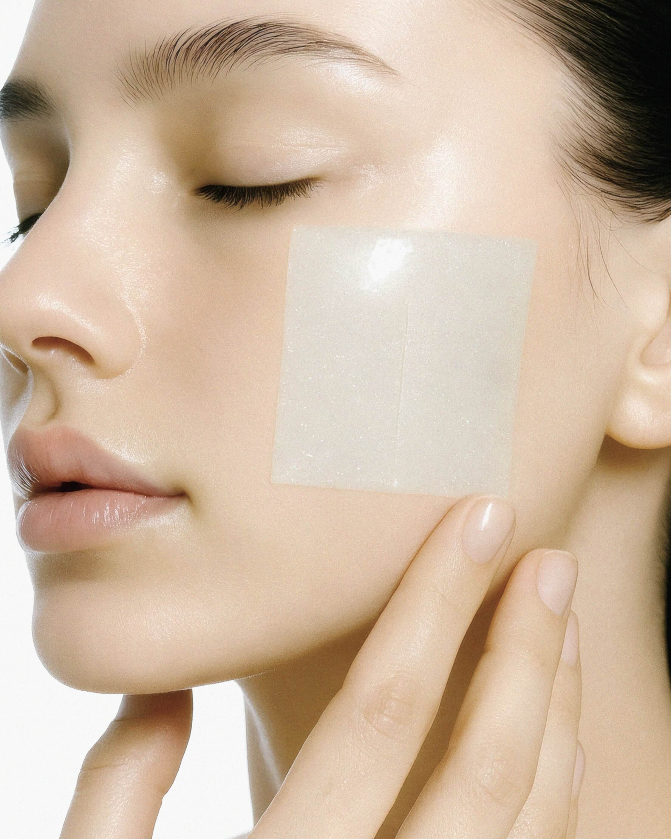 Close-up of a model applying a square, semi-transparent gel pad to her cheek, with one hand gently holding the pad in place.