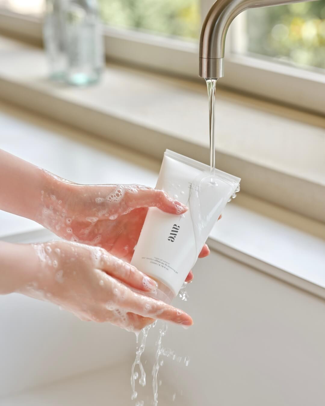 Close-up of soapy hands washing an Awe gentle cleansing gel tube under running tap water.
