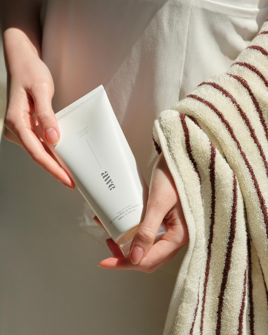 Woman holding an Awe natural cleansing gel tube next to a striped cotton towel.