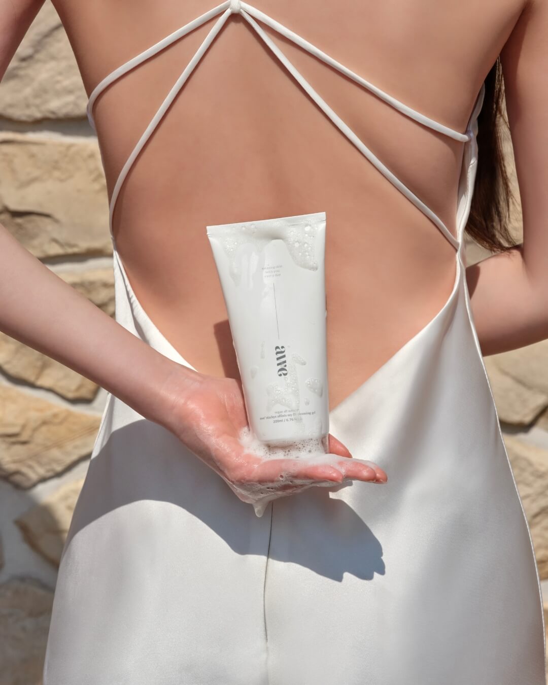 Woman in a white satin dress holding a foamy Awe cleansing gel tube behind her back.