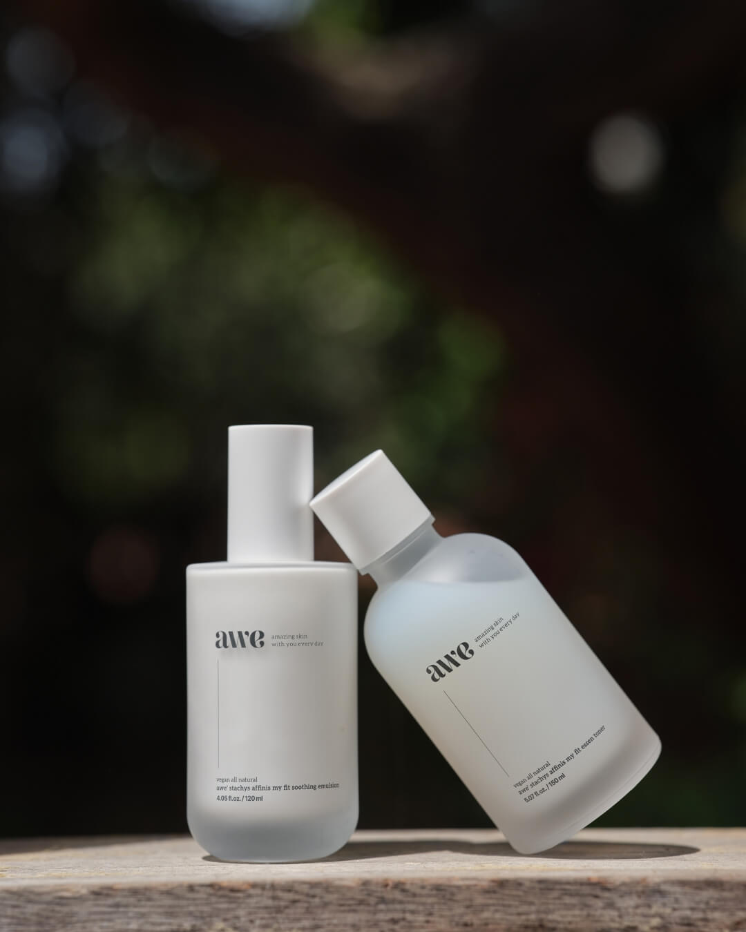 Awe facial essence toner and soothing emulsion bottles leaning together outdoors.