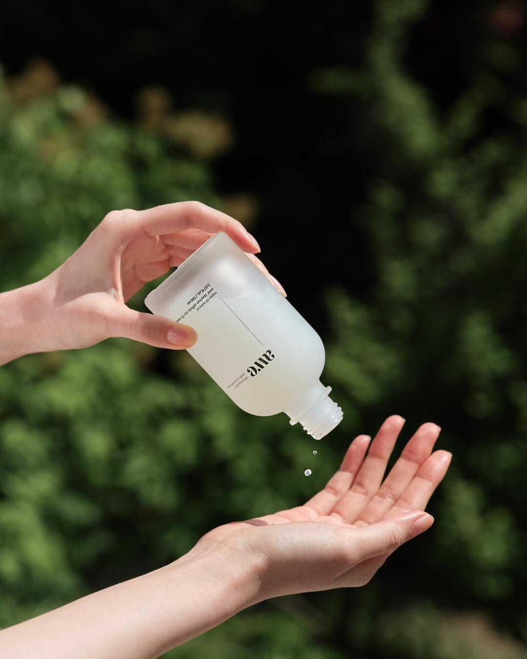 Hands pouring liquid from an Awe hydrating essence toner bottle against green leaves.