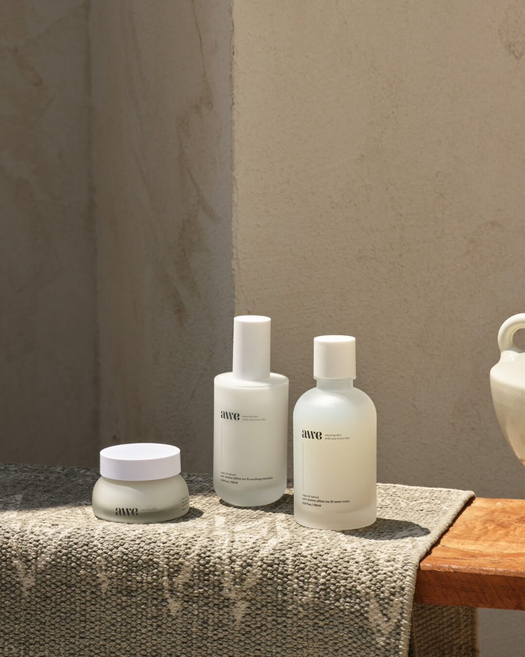 Awe essence toner, emulsion, and cream skincare routine setup on a sunny stone bench.