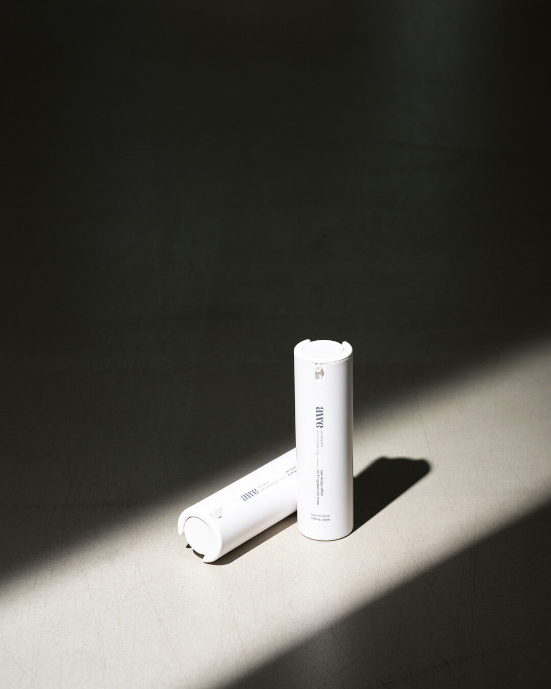 Two Awe signature eye cream bottles placed on the floor in a strong, dramatic beam of light.
