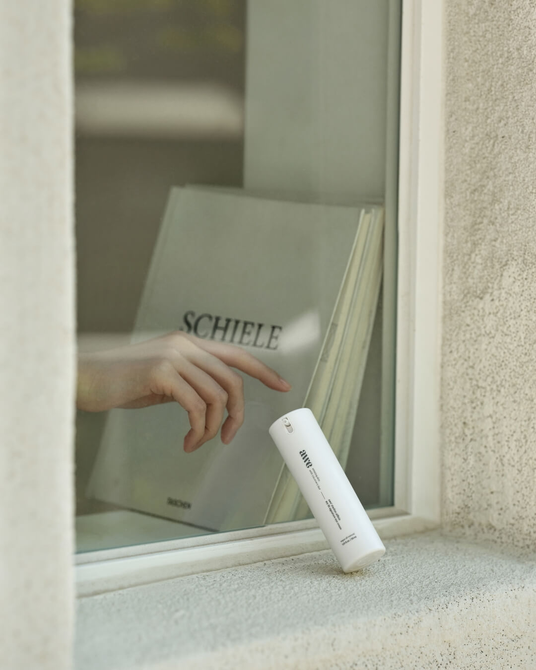 Awe hydrating eye cream bottle on a sunny window sill next to a book titled Schiele.