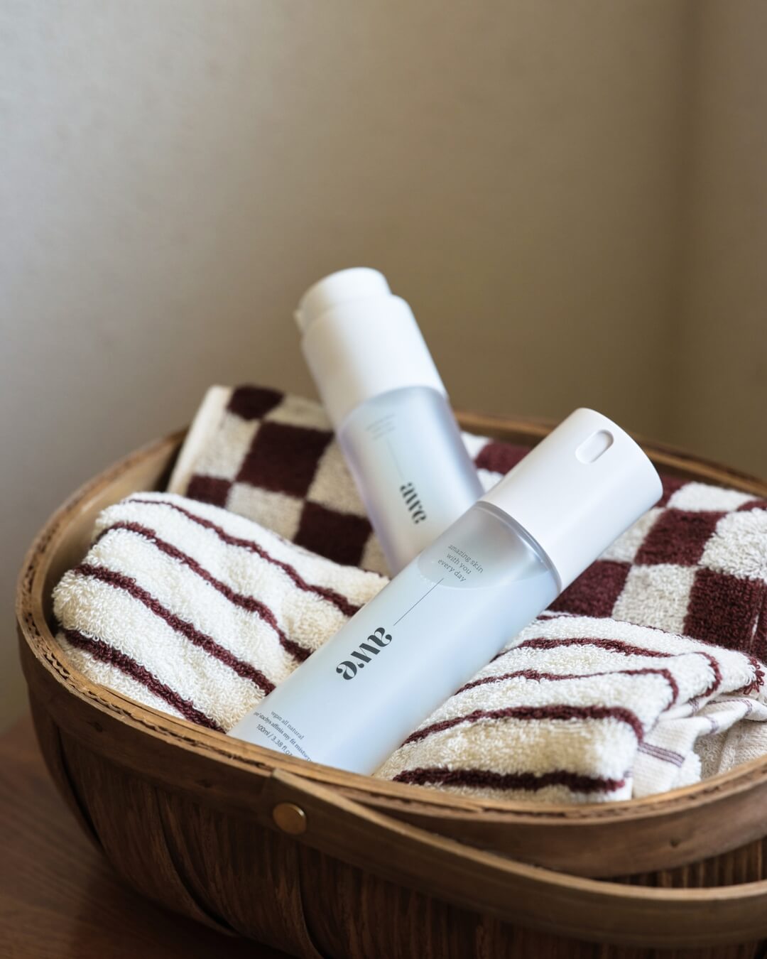 Two bottles of Awe firming mist serum placed inside a wooden basket with checkered towels.