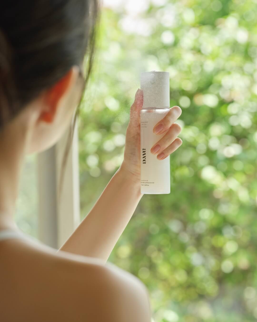 Woman spraying Awe firming mist serum bottle outdoors with lush green trees in the background.