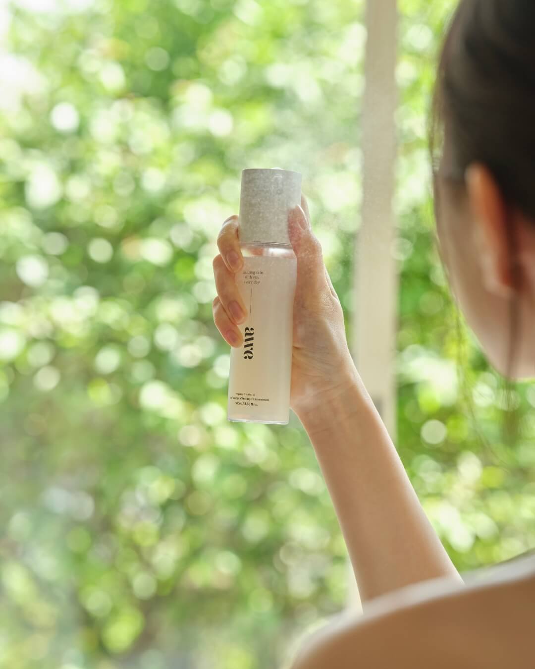 Close-up of a woman’s hand holding and spraying Awe skincare firming mist serum near a window.