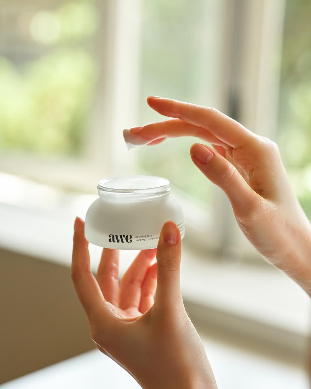 Woman dipping her finger into an open jar of Awe facial radiance cream near a bright window.
