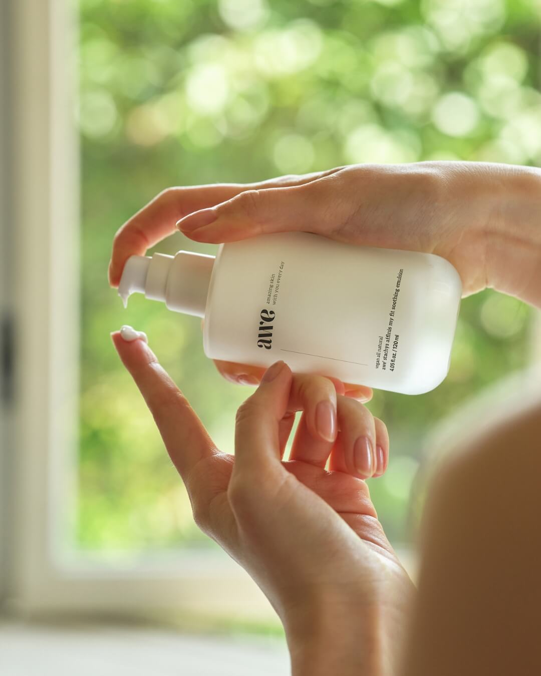 Close-up of a woman pumping white cream from an Awe daily soothing emulsion bottle onto her finger.