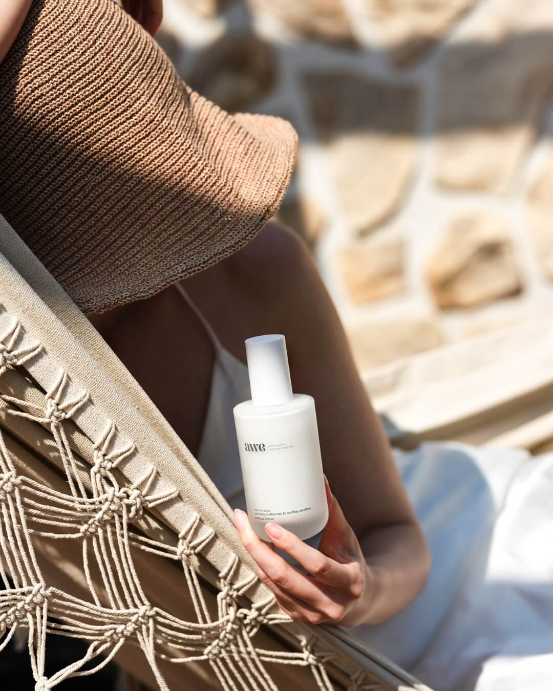 Woman in a straw hat sitting in a macrame hammock and holding an Awe facial soothing emulsion bottle.