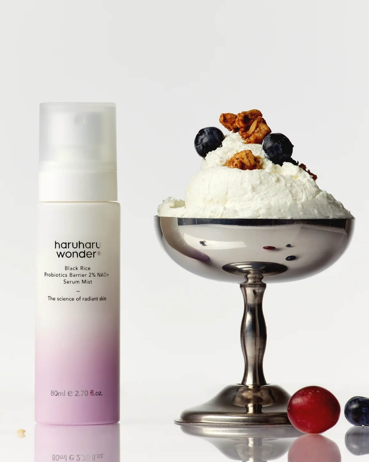 The serum mist bottle standing next to a silver dessert bowl filled with white cream, blueberries, and granola on a bright white surface.