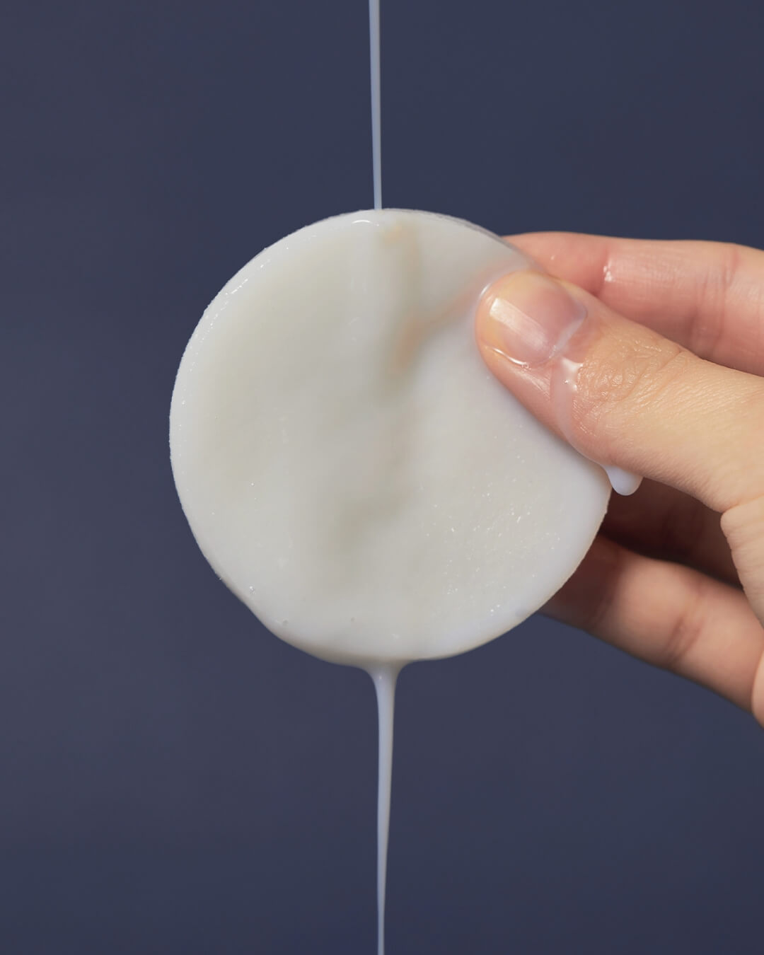 A hand holding a round koy Flow Intensive Cream Pad dripping with rich, milky moisturizing essence.