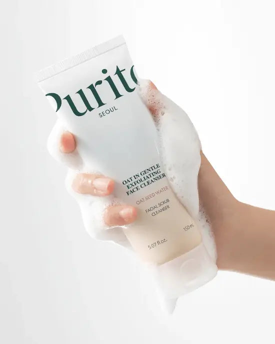 A hand gently grips the Purito Oat-in Gentle Exfoliating Face Cleanser tube, which is covered in a soft, white cleansing foam, emphasizing the product's mild foaming action and 'OAT SEED WATER' ingredient.